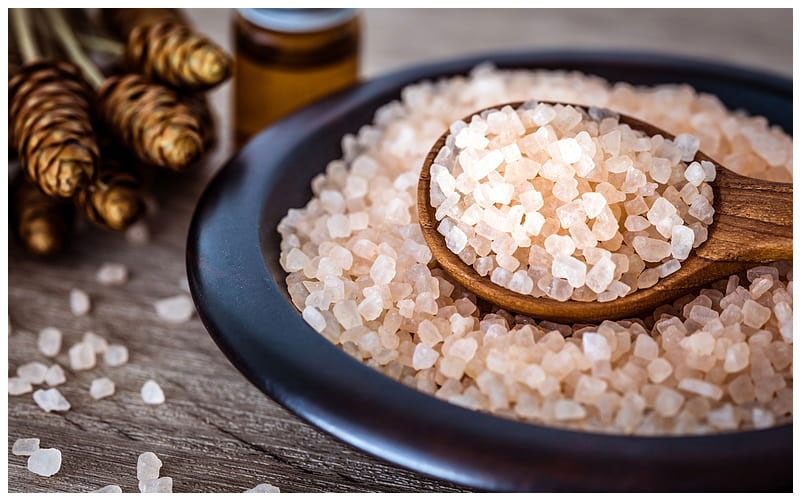 Light-pink coarse Himalayan salt crystals piled in a dark ceramic bowl with a wooden spoon